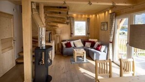 Een knus interieur van een blokhut met een houten trap, een bank met kussens, een tafel en een groot raam.