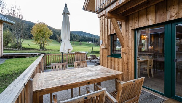 Een houten terras met een tafel en stoelen, een overdekte parasol en uitzicht op een groen landschap met heuvels op de achtergrond.