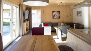 Ett modernt vardagsrum med trägolv, en soffa, ett soffbord och ett matbord. Det finns ett stort fönster med gardiner, en klocka på väggen och en liten hylla med diverse föremål.