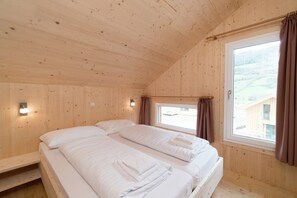 A wooden cabin room with two beds, a window with curtains, and a wall-mounted light.