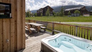 Una terraza de madera con un jacuzzi, mesa de picnic y bancos, con vistas a un paisaje montañoso.
