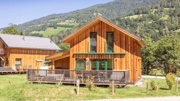 Una cabaña de madera con balcón, ubicada en un paisaje rural con colinas al fondo.