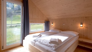 Une chambre dans une cabane en bois, avec une grande fenêtre, un lit recouvert de draps blancs et une vue sur un paysage verdoyant.