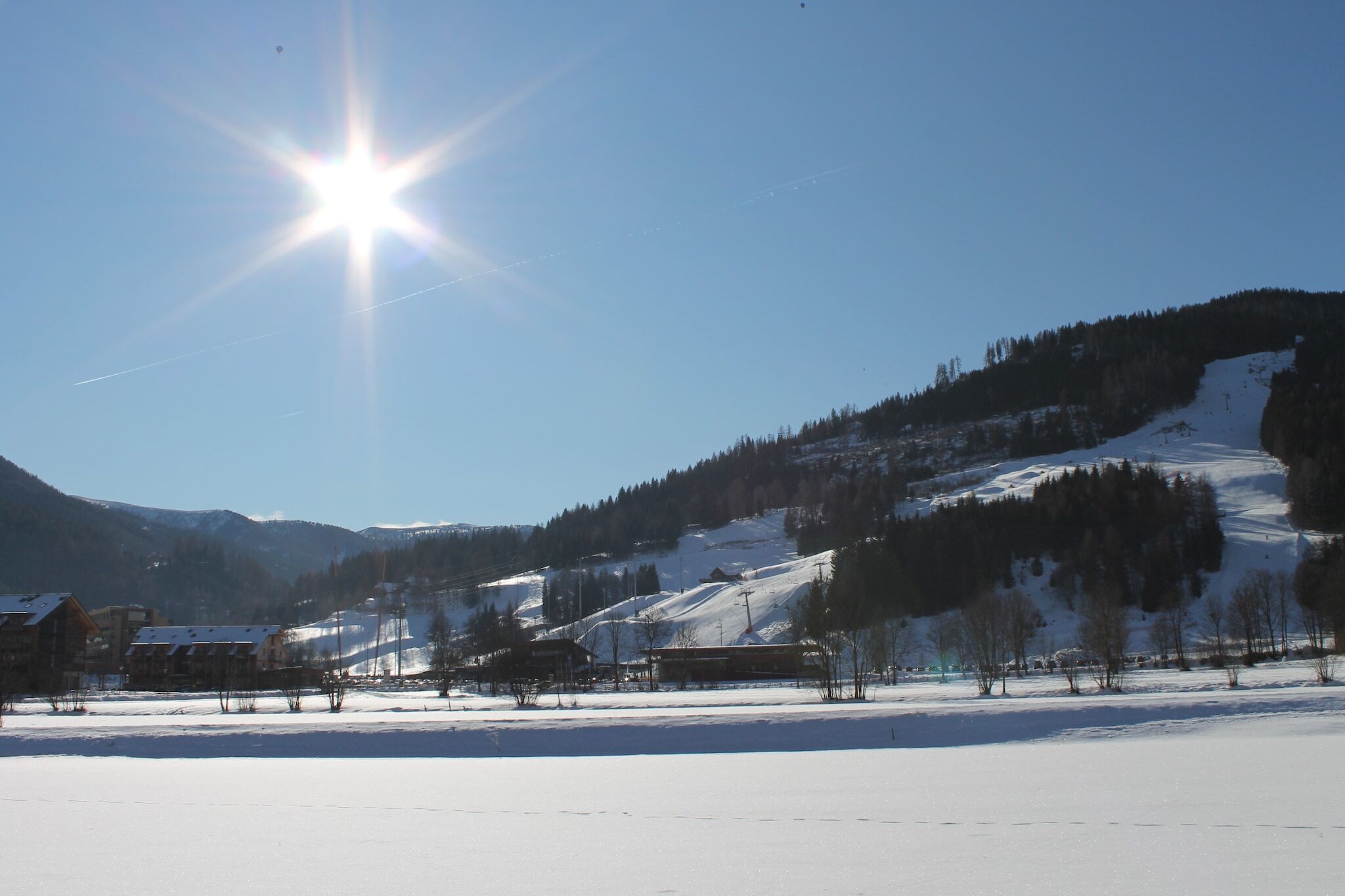 A snow-covered landscape with a clear sky, a sun shining brightly, and a mountainous backdrop.