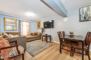 Una sala de estar con sofá, una mesa de comedor con sillas, un televisor montado en la pared y un cuadro.