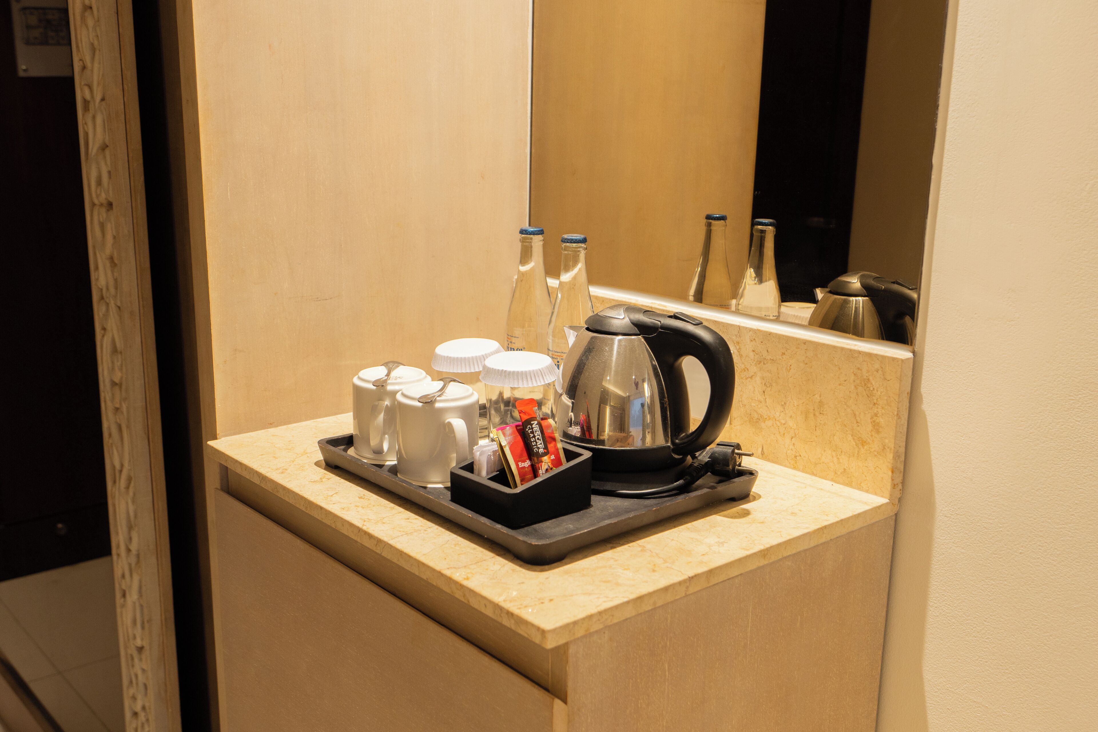 A hotel room's minibar with a tray containing cups, bottles, and a kettle.