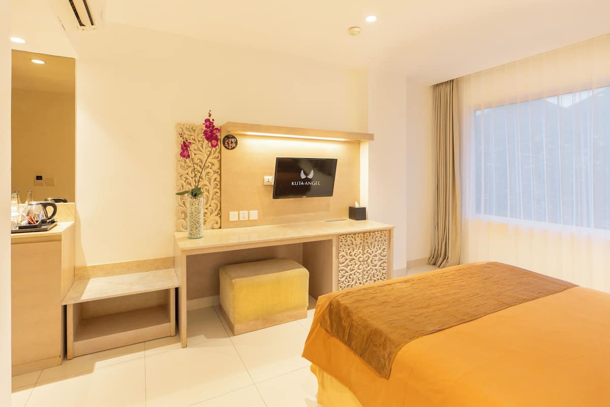 A hotel room with a bed, a desk with a TV, a small table with a vase, and a window with curtains.