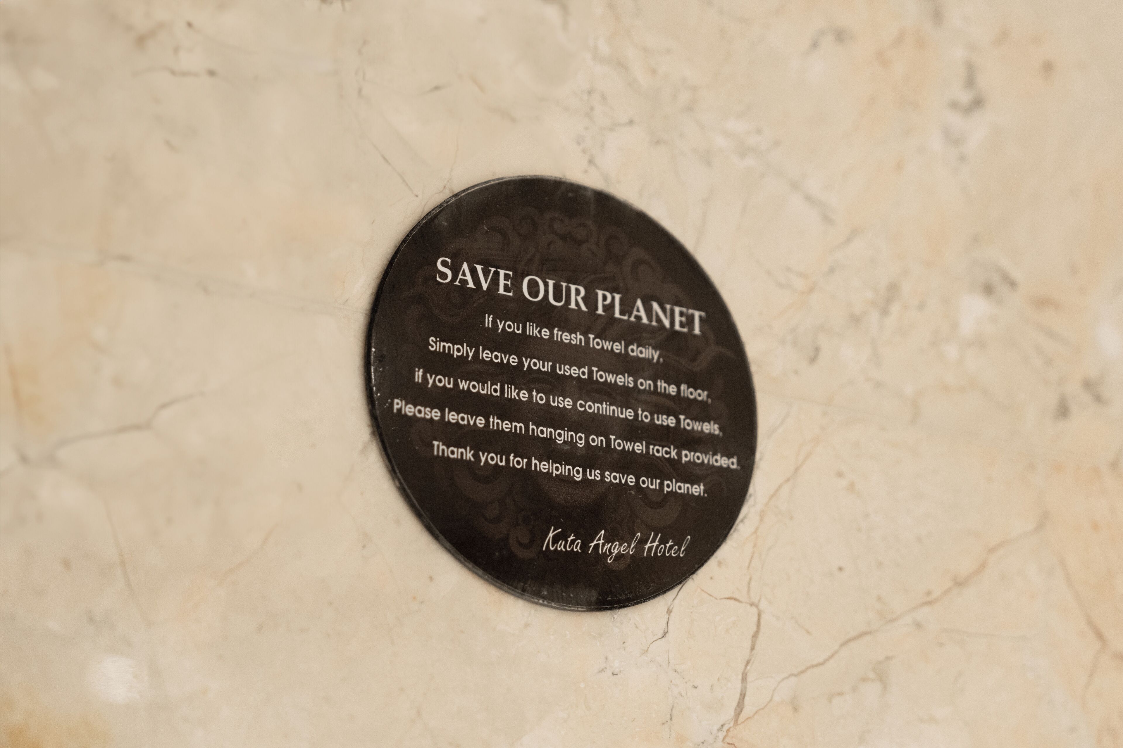 Ein rundes Schild mit dem Text „UNSERN PLANETEN RETTEN“ und Anweisungen zur Nutzung von Handtüchern, an einer Marmorwand befestigt.