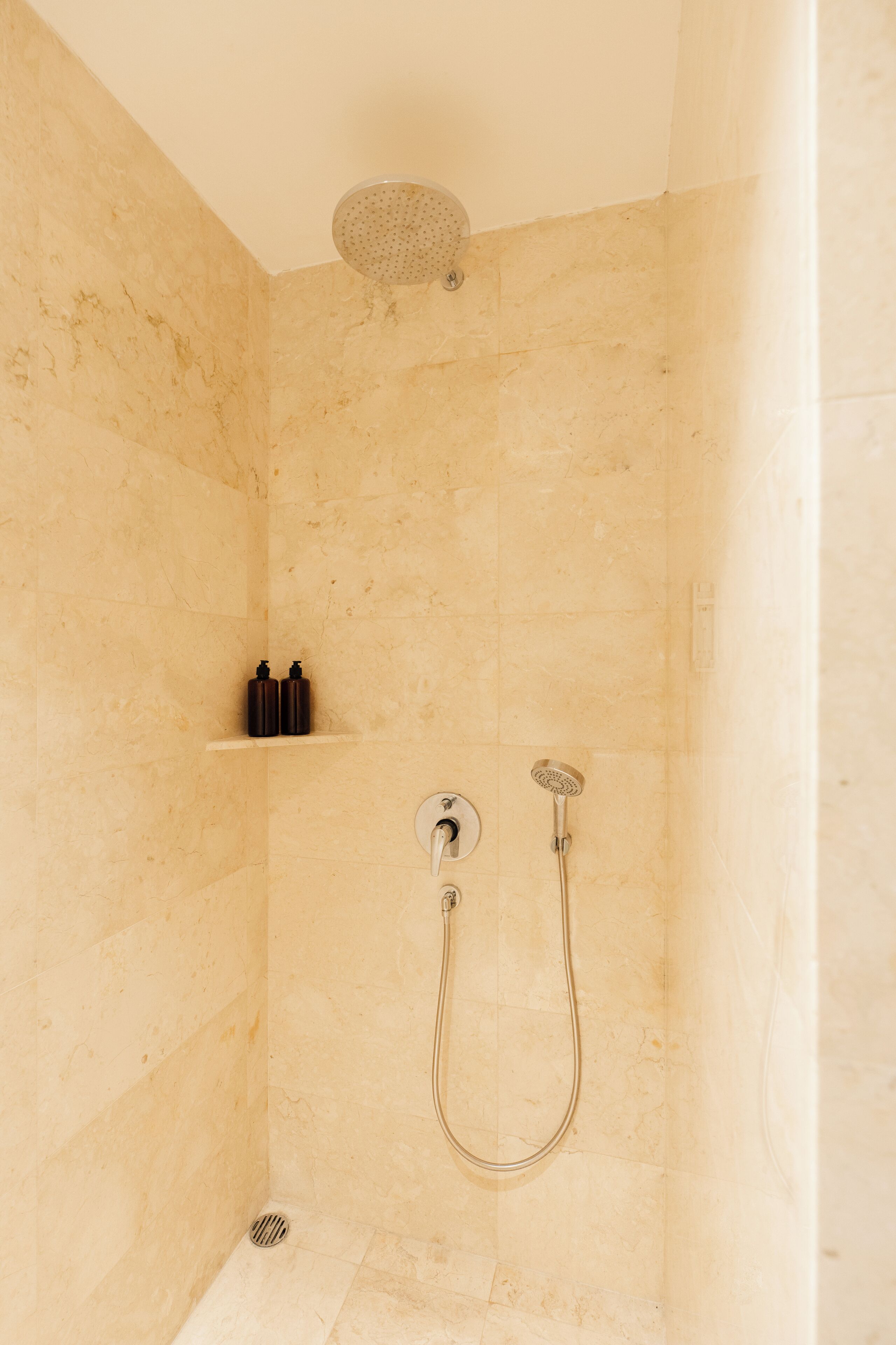 Ducha con cabezal de ducha de mano y grifo empotrado. En la pared, hay un estante con dos botellas.