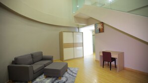 Ein modernes Wohnzimmer mit einem grauen Sofa, einem kleinen Hocker und einem Holztisch mit einem Stuhl. Eine gläserne Treppe führt zu einer oberen Ebene.