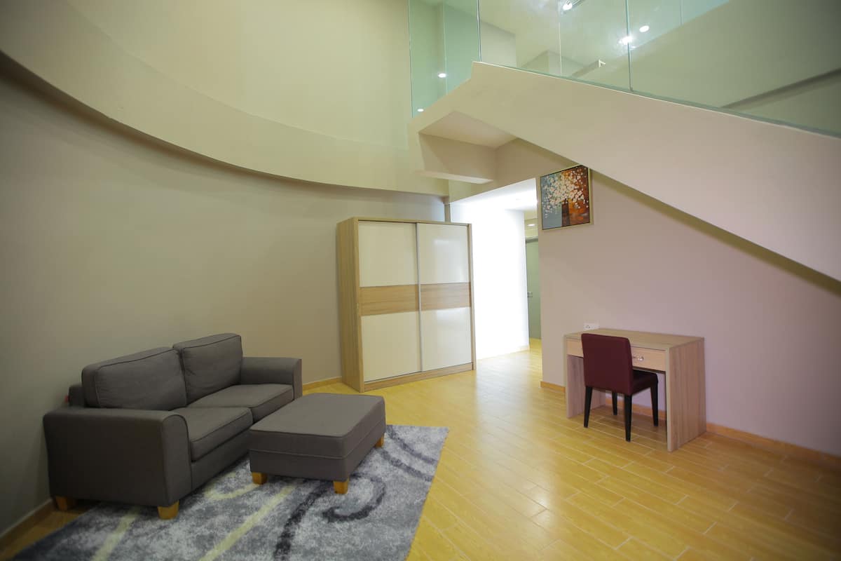 Ein modernes Wohnzimmer mit einem grauen Sofa, einem kleinen Hocker und einem Holztisch mit einem Stuhl. Eine gläserne Treppe führt zu einer oberen Ebene.