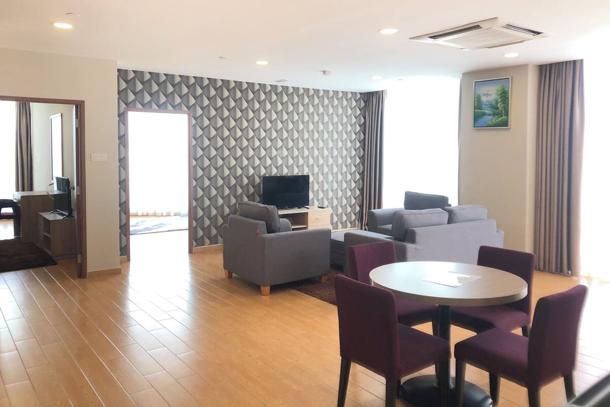 Ein modernes Wohnzimmer mit einem Flachbildfernseher, grauen Sofas, einem runden Esstisch mit violetten Stühlen und einer Wand mit Muster.
