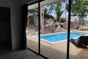A modern room with a large sliding glass door leading to an outdoor swimming pool area with rocks and trees. - Oikos, an open space toward the sky, toward yourself, and toward others (Tambon Ban Tai)