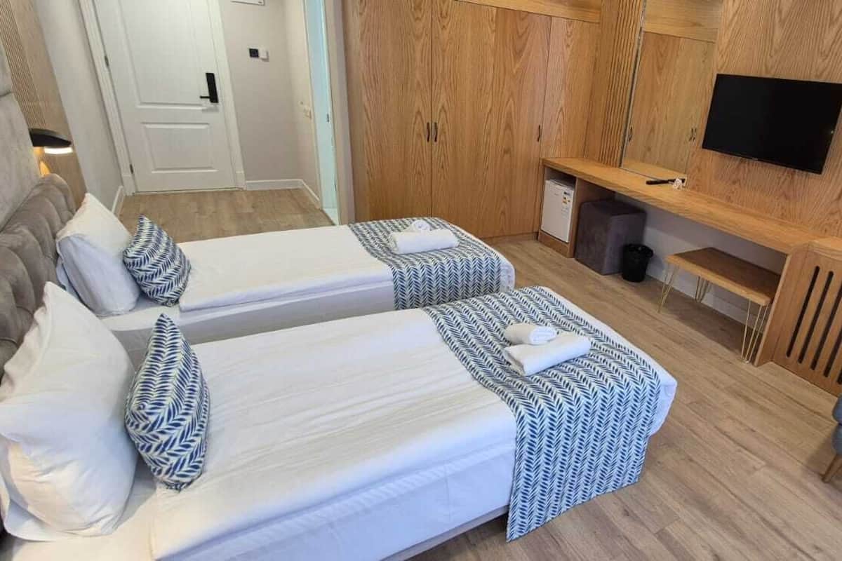 Habitación de hotel con dos camas, un sillón gris, un mueble de televisión de madera y una mesita con artículos de aseo.