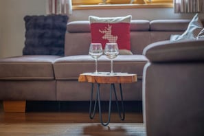 A round wooden table with metal legs, two wine glasses on it, and a red pillow with a reindeer design on a sofa in the background.
