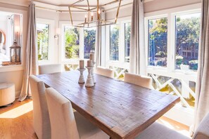 A dining area with a wooden table, white upholstered chairs, and a view of greenery through large windows.