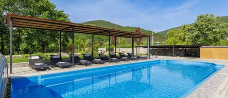 A swimming pool with lounge chairs and a pergola, surrounded by greenery and hills.