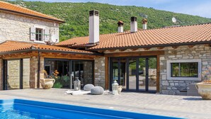 Un edificio de piedra con piscina, ventanas amplias y montañas al fondo.