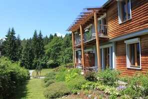 Une cabane en bois dotée d’un balcon, entourée d’une végétation luxuriante et bordée par une forêt.