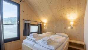 A wooden cabin bedroom with a bed, two bedside tables, a window with curtains, and a view of a lake.