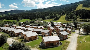 Eine Gruppe Holzhütten mit Satteldächern, angeordnet in einer kleinen Siedlung, umgeben von üppigen grünen Feldern und bewaldeten Hügeln.