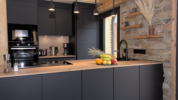 A modern kitchen with dark cabinetry, a wooden countertop, and a stone accent wall. - Attractive chalet with sauna (Sankt Georgen am Kresichberg)