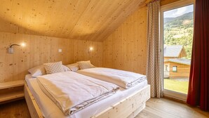 Un dormitorio de cabaña de madera con dos camas, una ventana con cortinas y vistas a una casa y un paisaje.