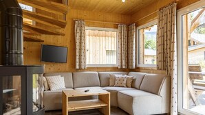 Interior acogedor de una cabaña con un sofá modular, una mesa de madera, una chimenea y ventanas grandes con cortinas.