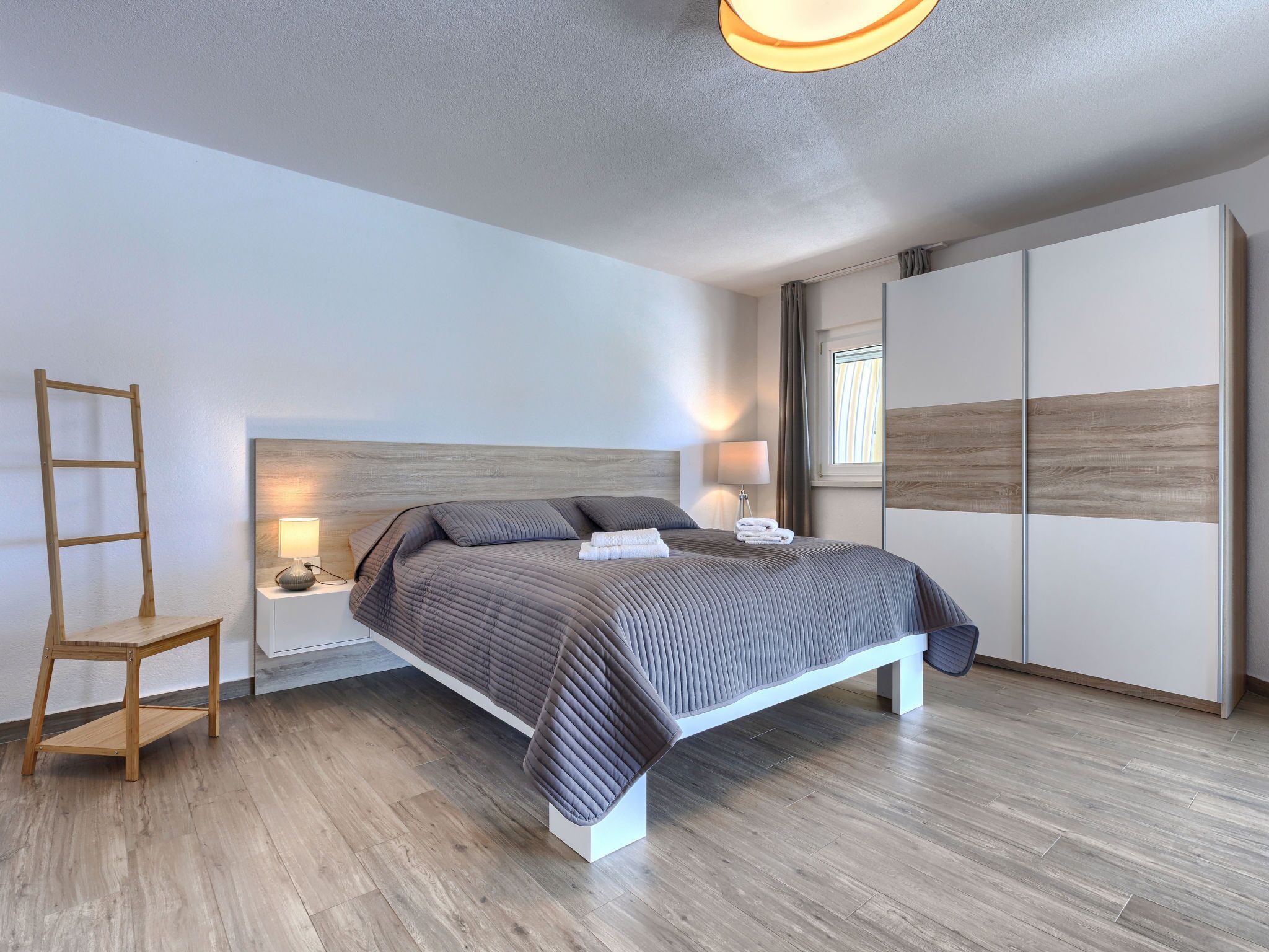 Quarto moderno com cama de madeira, mesas de cabeceira com luminárias, um guarda-roupa e uma escada encostada na parede.