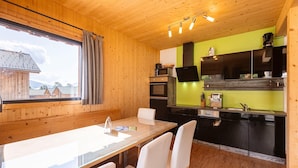Un interior de madera con una mesa de comedor y sillas, una cocina con armarios negros y una pared verde, y una ventana con cortinas.
