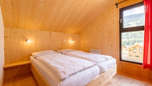 Una habitación de madera con una cama, un ventanal con cortina roja y vista a una casa.