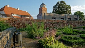 Een historisch gebouw met een bakstenen muur en een toren, omringd door een tuin met diverse planten en een bankje.
