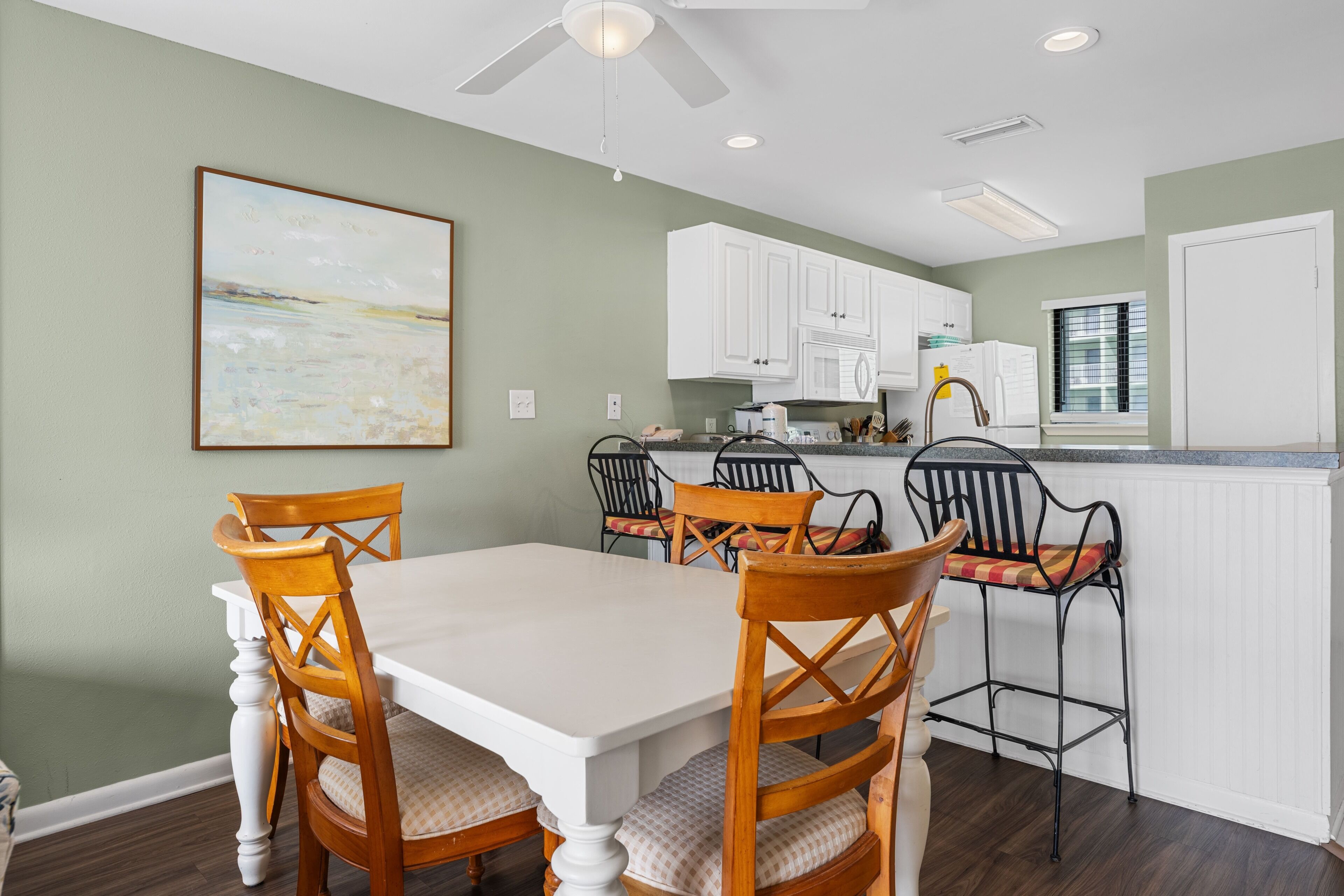 A dining area with a white table and wooden chairs, a kitchen with white cabinets, and a ceiling fan.