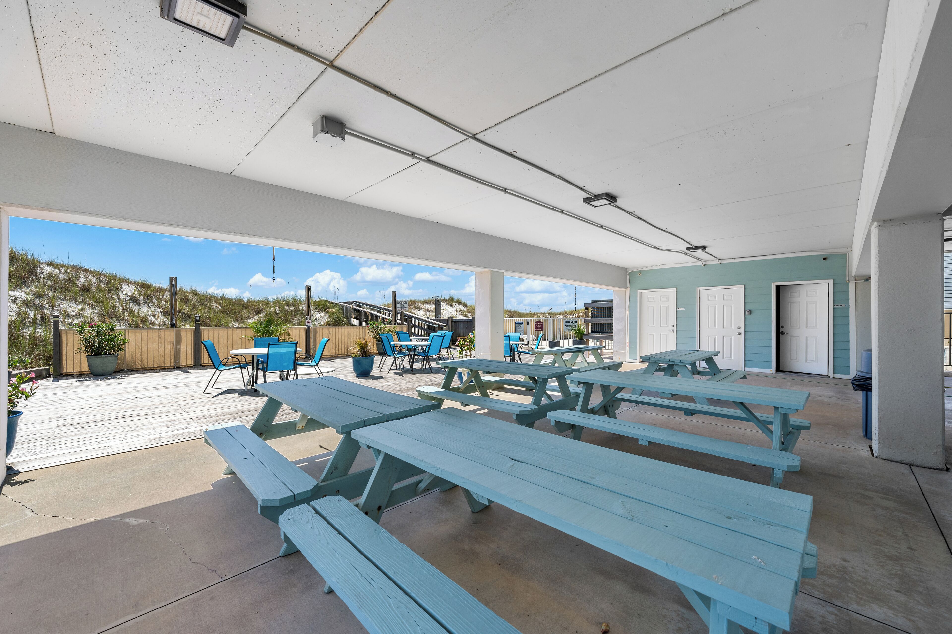 Un patio al aire libre con mesas de picnic y sillas azules, una terraza de madera y vista a la playa.