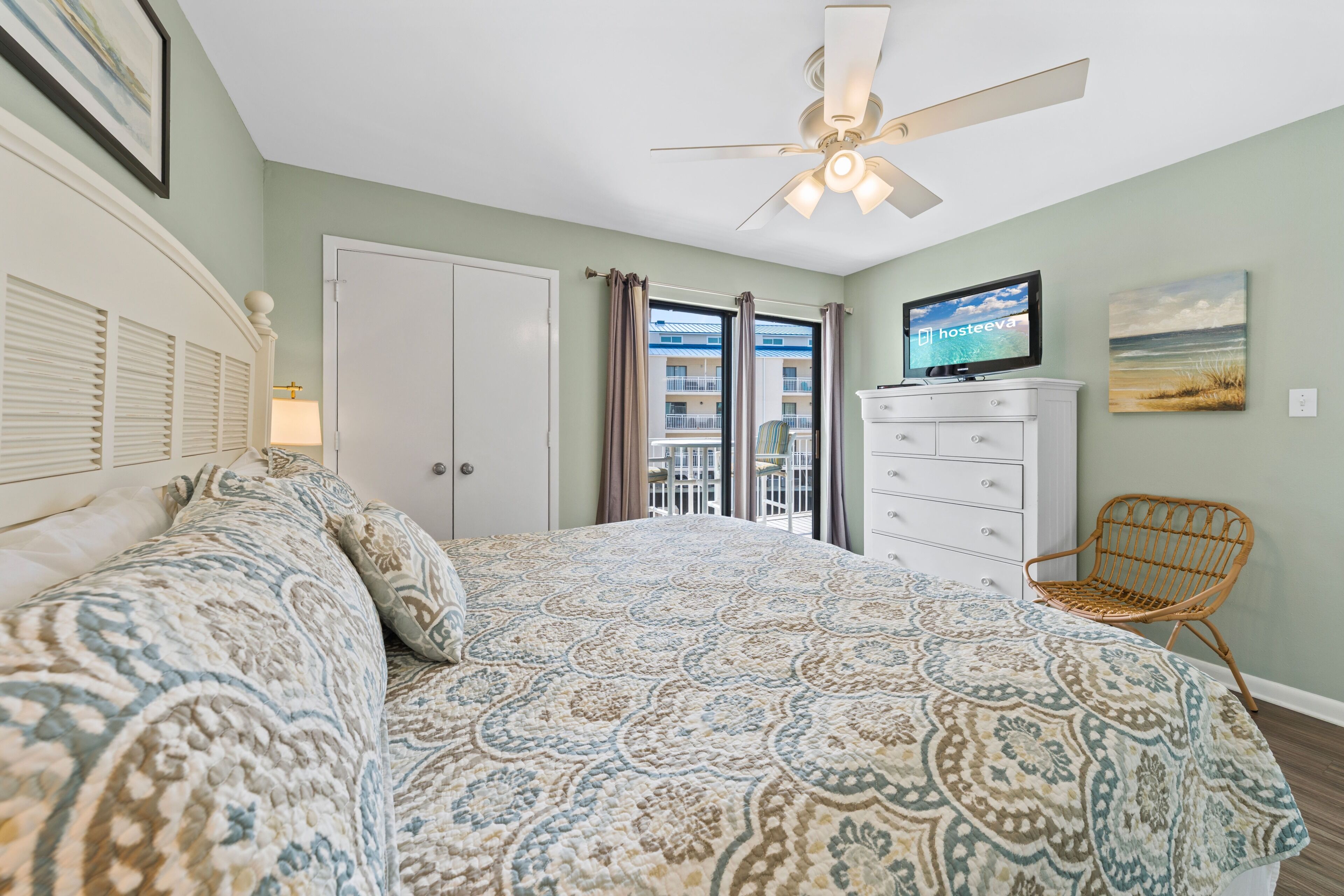 Un dormitorio con cama, ventilador de techo, televisión, cómoda, silla y balcón con vistas a edificios.