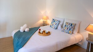 Una cama bien hecha con un edredón verde azulado, dos almohadas estampadas y una bandeja con cruasanes y tazas de café.