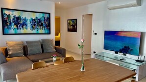 A modern living room with a sofa, a dining table, a vase with a flower, and a TV displaying a sunset scene. - B12a - 2 Bdr Serviced Apt with Pool Access, Karon Beach (Tambon Karon)