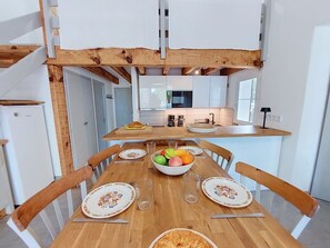 A dining area with a wooden table, chairs, and a fruit bowl. A kitchen is visible in the background with modern appliances. - Cosy apartment for 6 people with WIFI, TV, terrace and pets allowed (Mimizan)