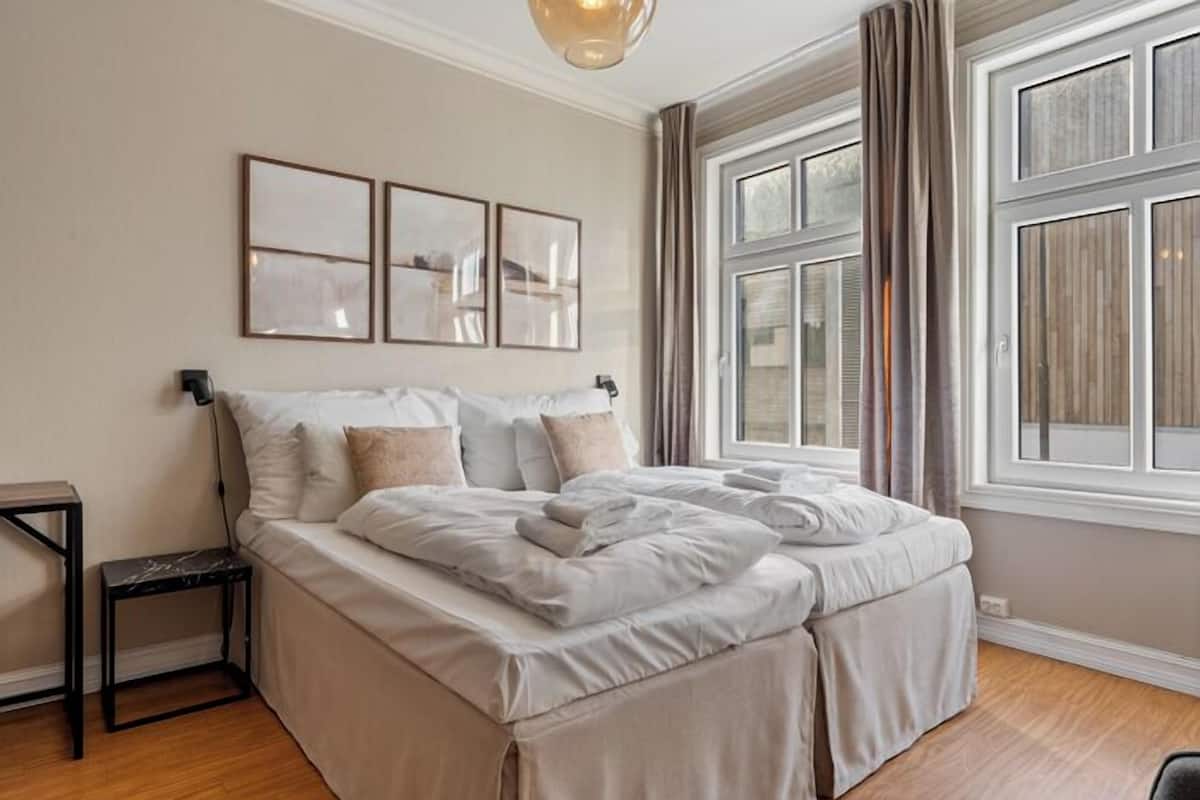 Une chambre à coucher bien rangée, avec un lit, des tables de chevet et une grande fenêtre avec des rideaux.