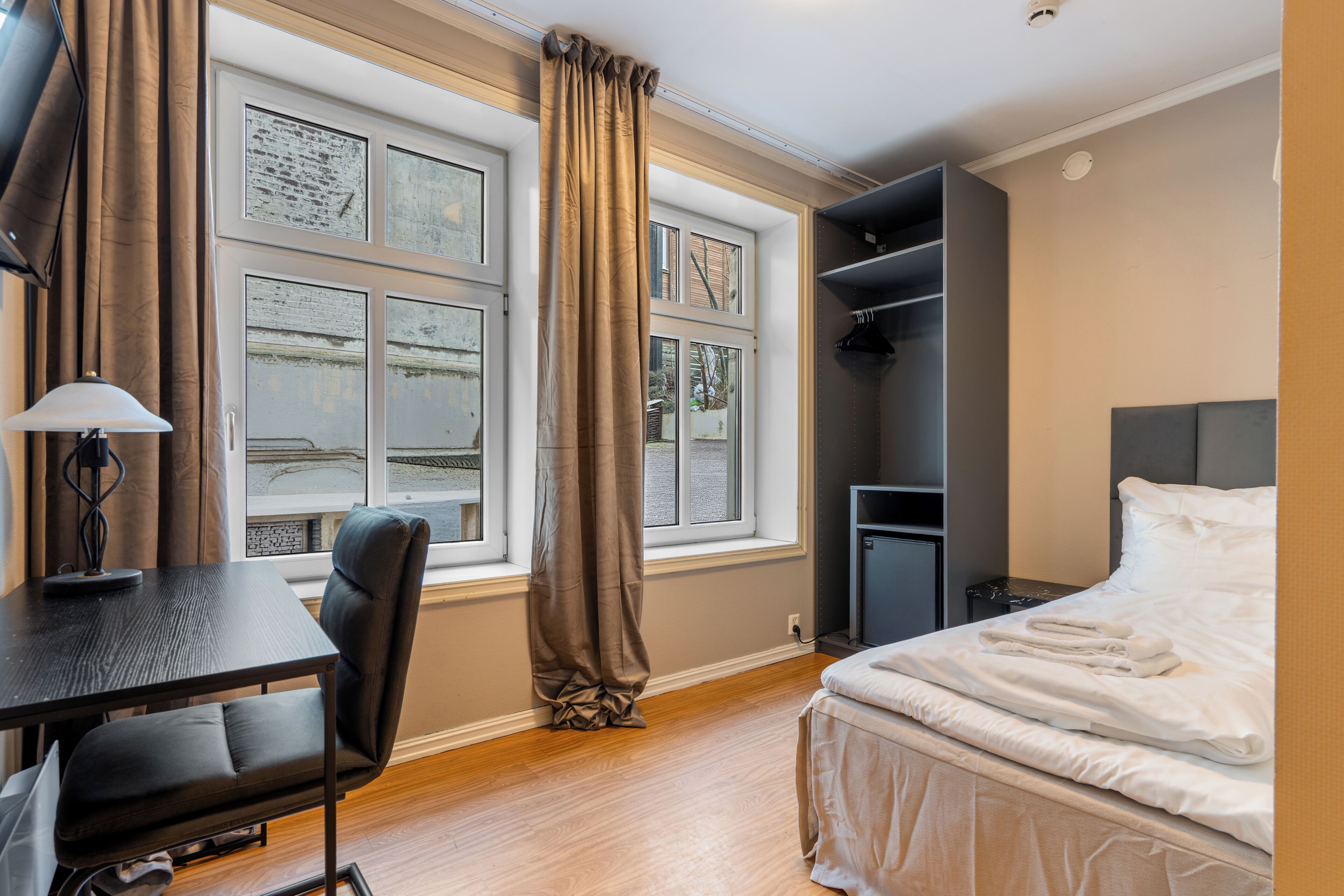 Una habitación de hotel con una cama, un escritorio con silla, un armario y una ventana con cortinas.