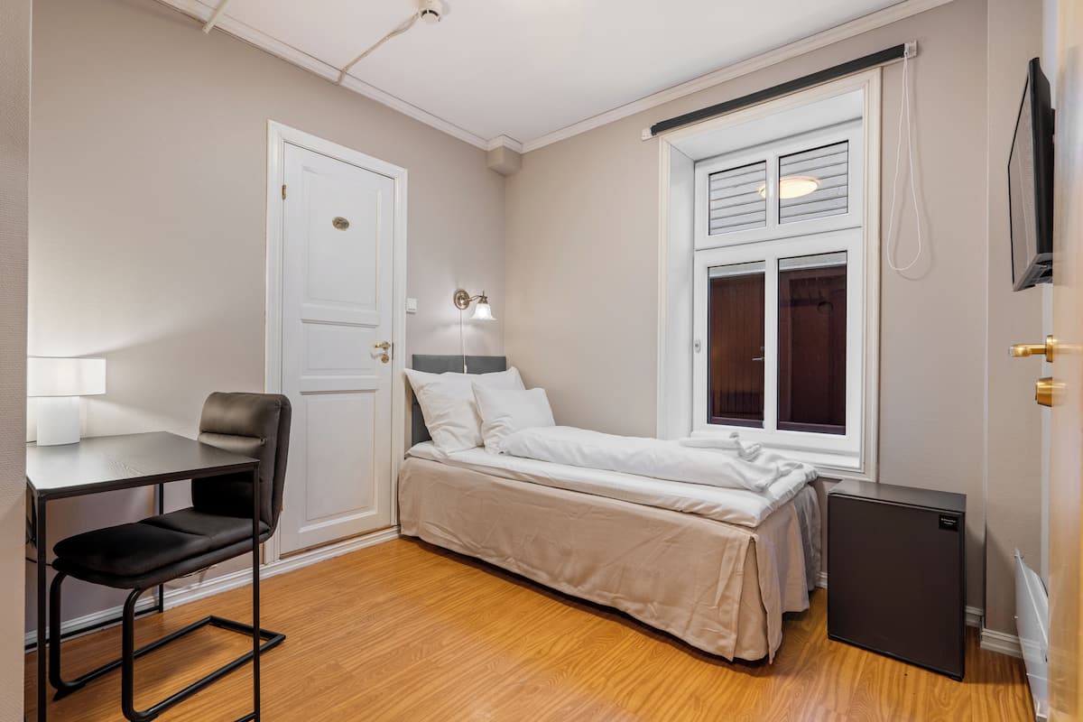 Une petite chambre à coucher, bien rangée, avec un lit, un bureau et une chaise, un petit réfrigérateur et une fenêtre avec des stores.