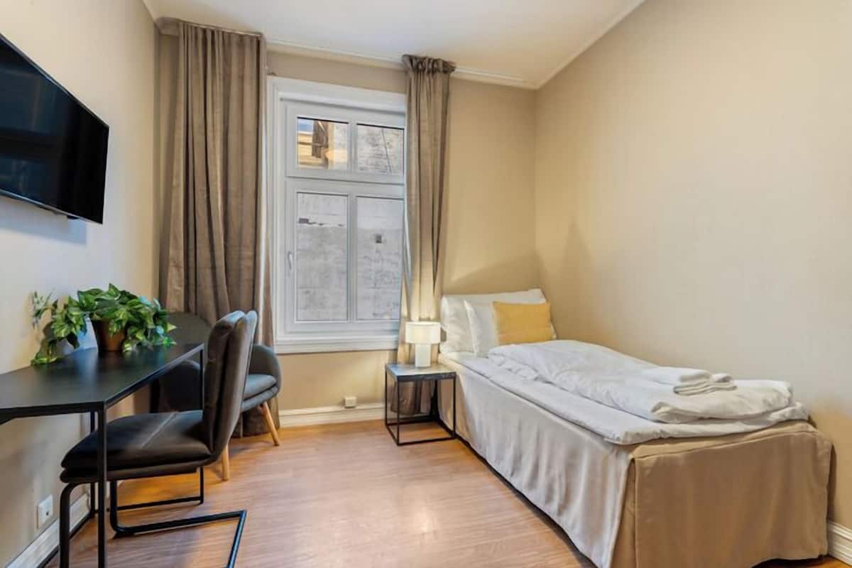 Une chambre d’hôtel de taille réduite, comprenant un lit, un bureau, une chaise, une télévision et une fenêtre avec des rideaux.