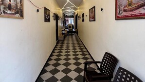 Un corridoio d'albergo con pavimento a scacchi, quadri alle pareti e una fila di sedie su un lato.