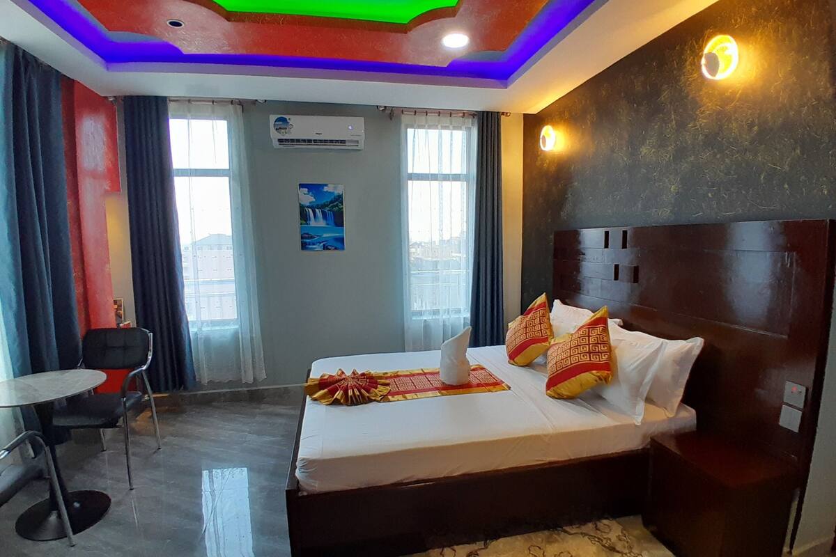 A hotel room with a bed, a desk, and a chair. There is a painting on the wall and an air conditioning unit.