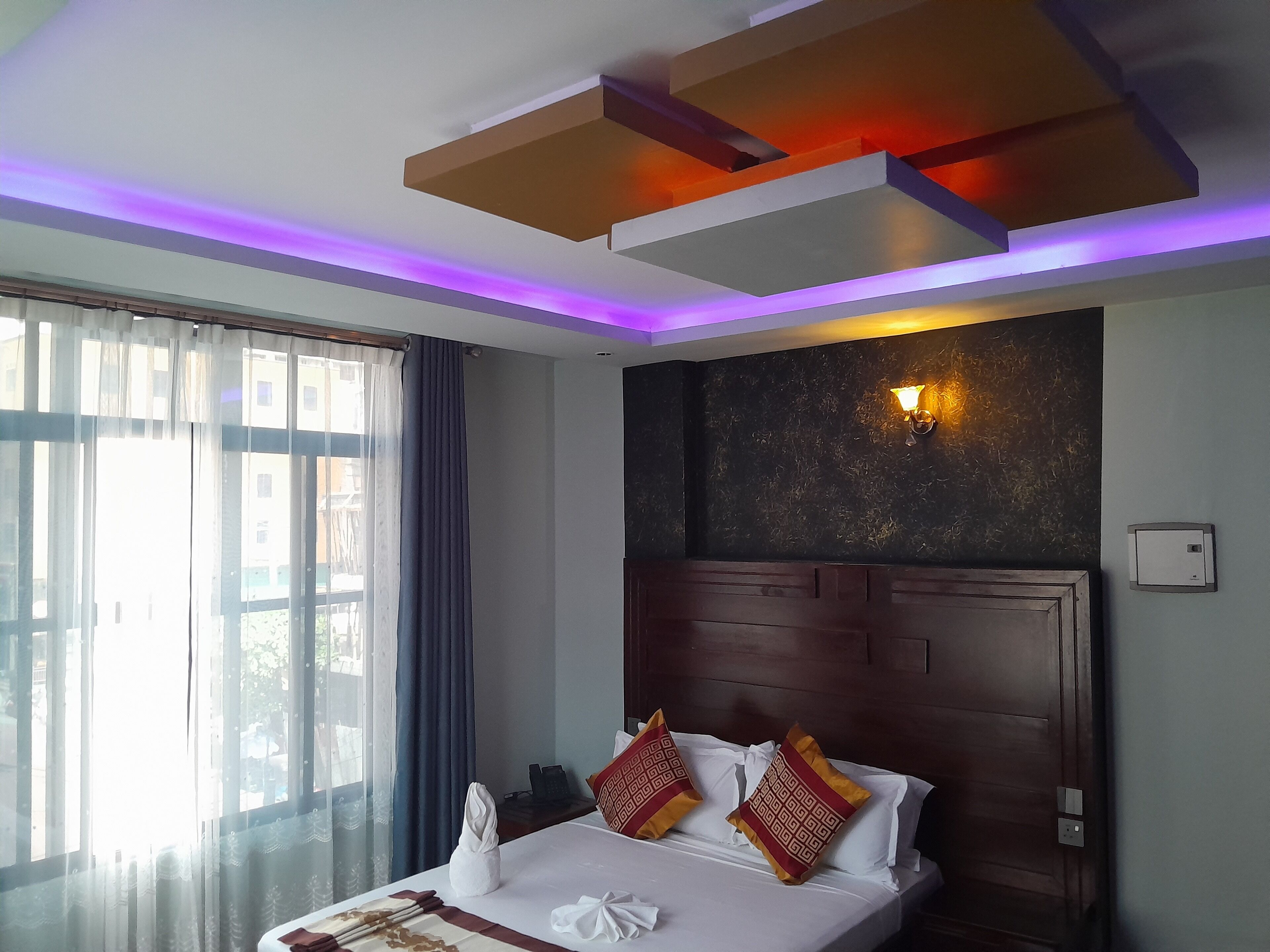 Une chambre d’hôtel avec un lit, une fenêtre avec des rideaux, un luminaire fixé au mur et un plafond doté d’un appareil d’éclairage.