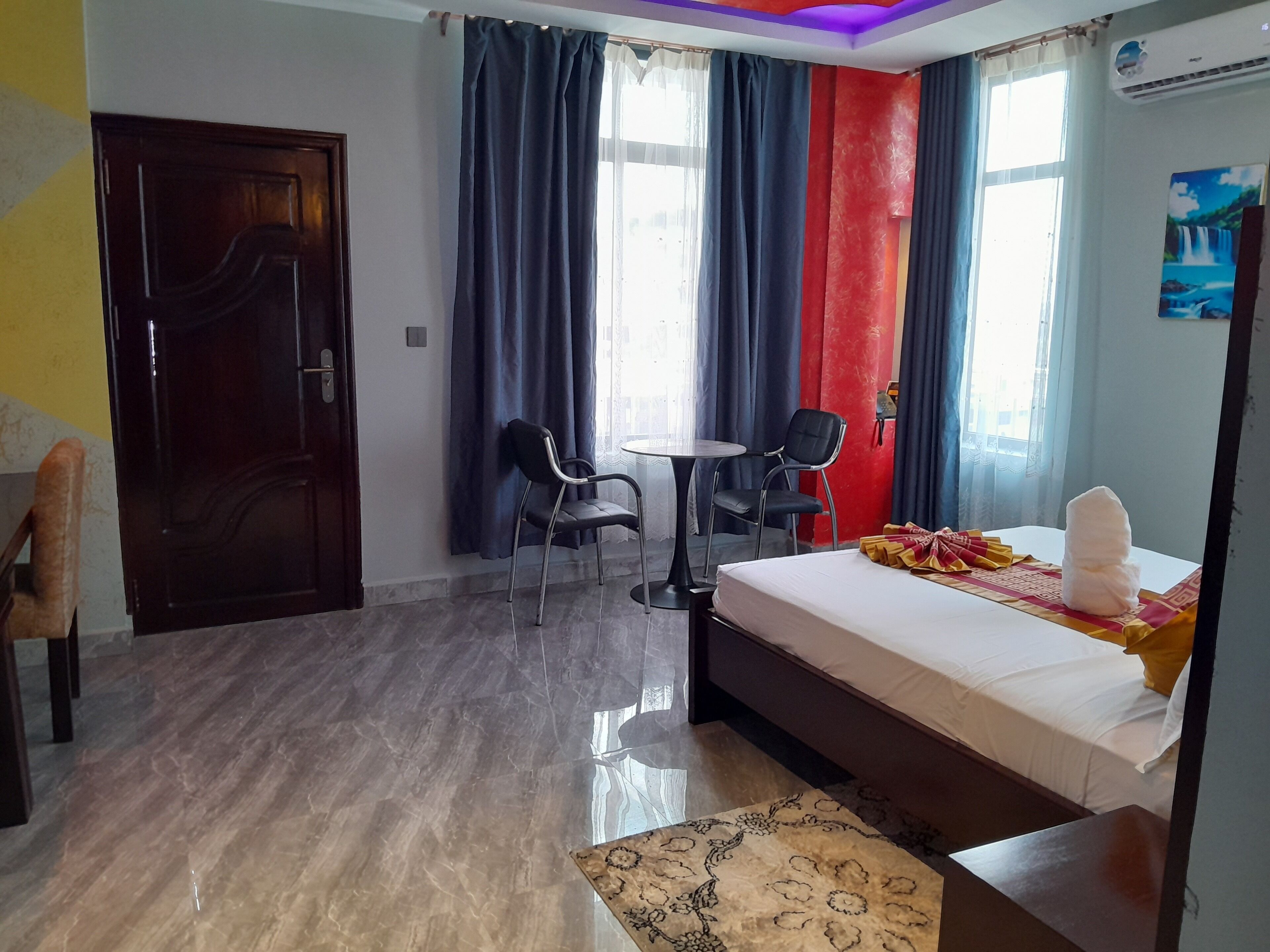 A hotel room with a bed, a desk, two chairs, a window with curtains, and a door.