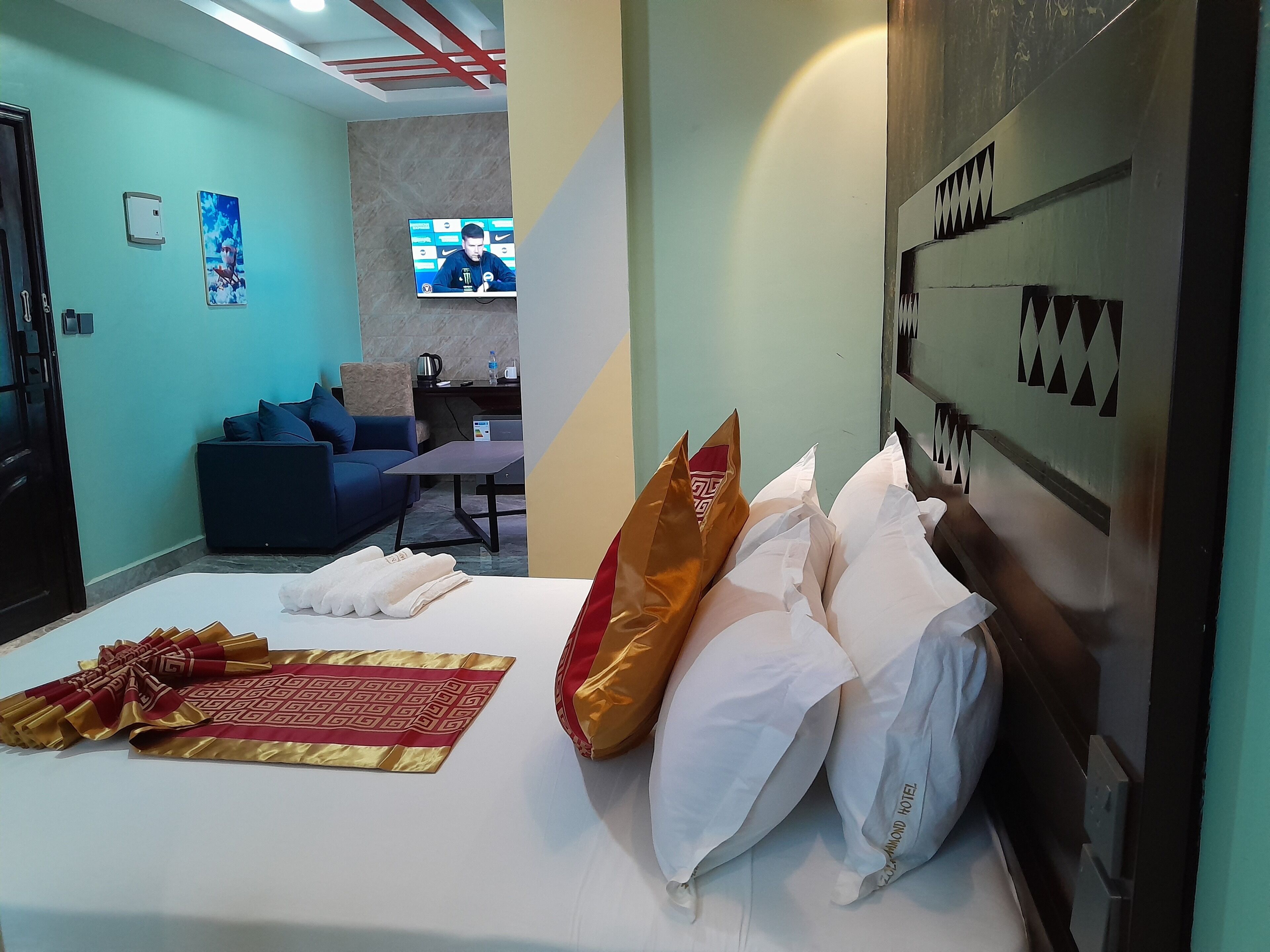 A hotel room with a bed, pillows, a blanket, a sofa, a TV, and a wall with a geometric pattern.