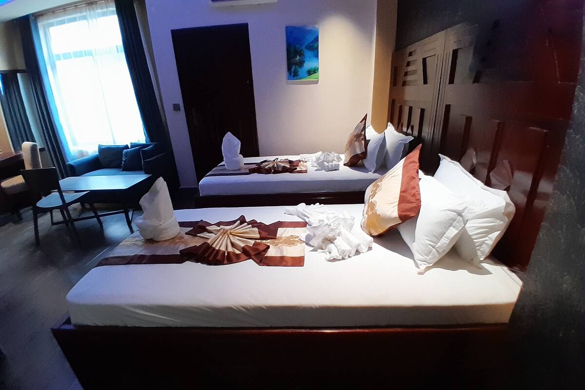 A hotel room with two beds, a dining table, and chairs. There is a window with curtains, a door, and a wall-mounted artwork.