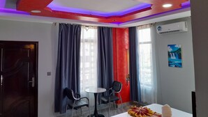 A hotel room with a red accent wall, two chairs, a small table, a bed, and an air conditioning unit. - MAZOLA DIAMOND HOTEL (Dar es Salaam)