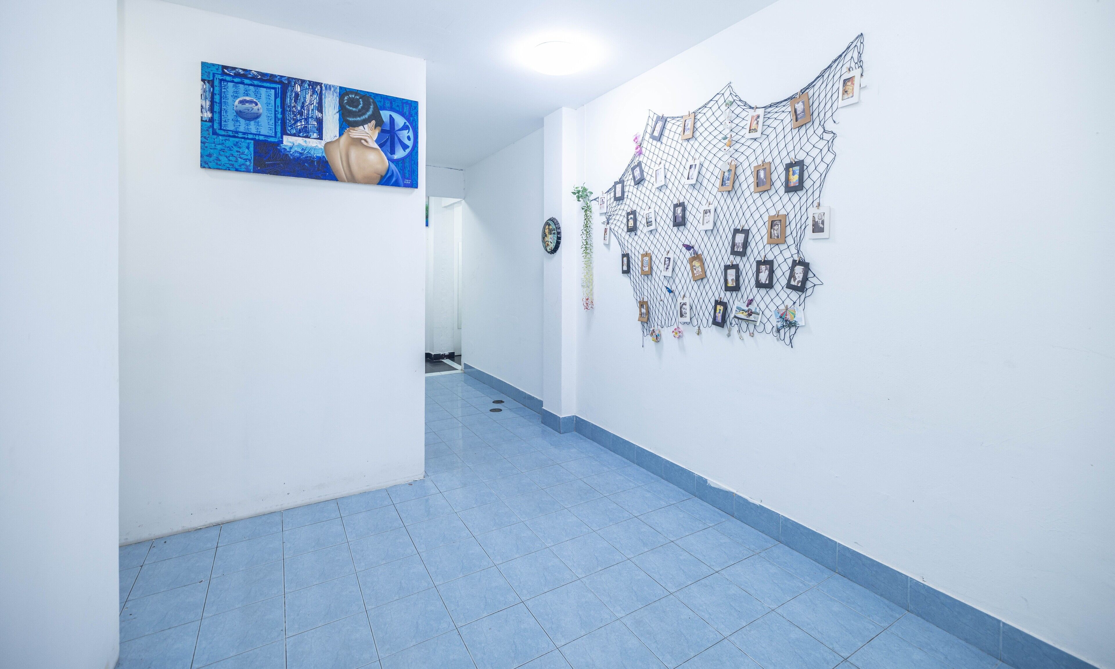 A hallway with a blue tiled floor, a large abstract painting on the left wall, and a wall-mounted photo display on the right.